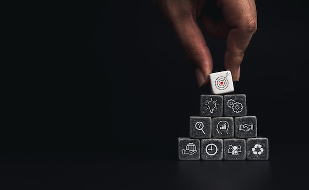 Business strategy with growth success process for leadership\
and teamwork concept. hand putting business target icon on dice\
cube blocks stack pyramid shape on dark background with copy\
space.