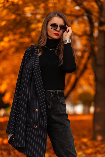 Business stylish woman in a fashionable black suit with a vintage blazer and a sweater straightens sunglasses in an autumn park with colored fall orange foliage