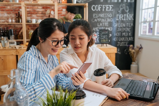 Business team partners working on laptop computer at coffee bar. two young girls colleagues looking at mobile phone together in modern cafe store. barista in apron prepare customer order in counter