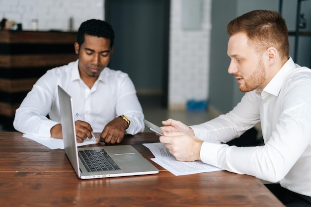 Business team van twee multi-etnische zakenlieden mensen in formalwear zitten samen op kantoor met laptop, papieren documenten, digitale tablet en bespreken nieuw project of bedrijfsidee.