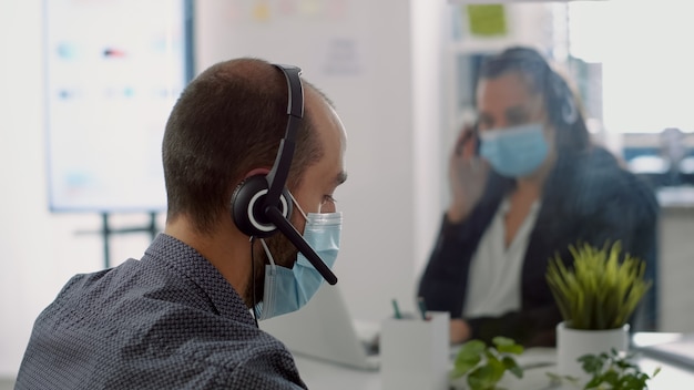 Team aziendale che indossa maschere protettive e cuffie, lavorando al computer durante l'epidemia globale di covid19. i colleghi mantengono il distanziamento sociale per prevenire la malattia da virus