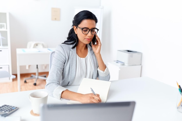 business, technology, communication and people concept - businesswoman or secretary calling on smartphone and writing to notebook at office