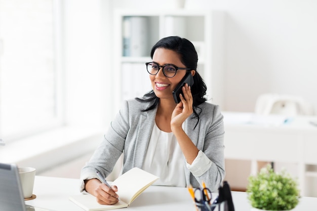 Photo business, technology, communication and people concept - happy smiling businesswoman or secretary with notebook calling on smartphone at office