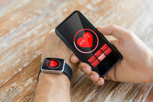 business, technology, health care and people concept - close up of male hand holding smart phone and wearing smart watch showing red heart beat icon on screen