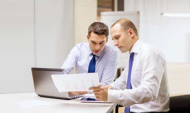 business, technology and office concept - two businessmen with laptop, tablet pc computer and papers having discussion in office