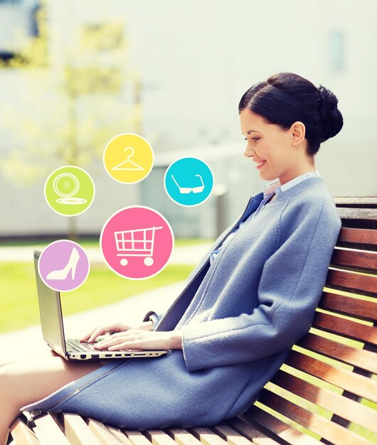 business, technology, online shopping and people concept - young smiling woman with laptop computer and internet icons sitting on bench in city