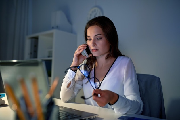 business, technology, overwork, deadline and people concept - woman with laptop calling on smartphone at night office