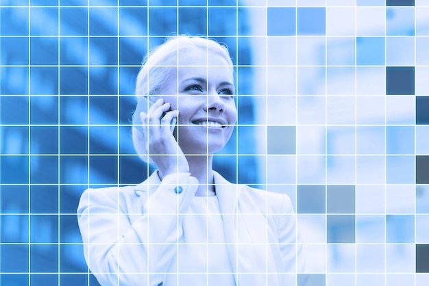 Photo business, technology and people concept - smiling businesswoman with smartphone talking over office building and blue grid background