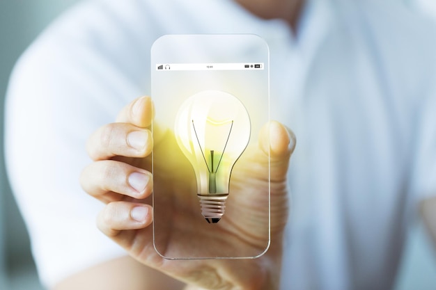 写真 ビジネス、テクノロジー、スタートアップ、アイデア、人のコンセプト – 電球のアイコンが付いた透明なスマートフォンを持ち、見せる男性の手の接写