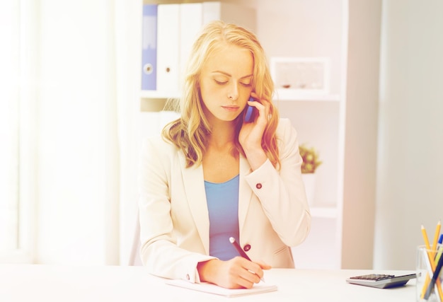 business, tecnology, communication and people concept - businesswoman with pen and notebook calling on smartphone at office