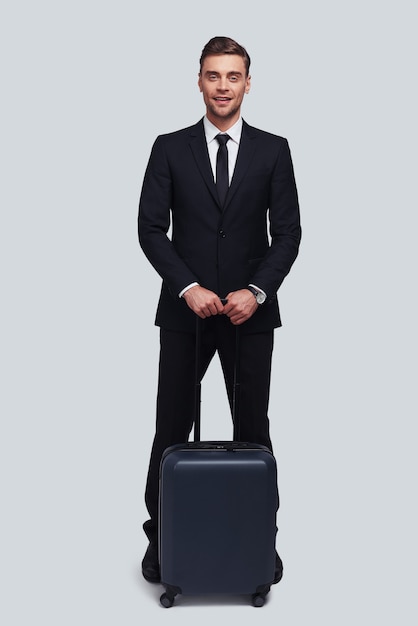 Business travel. Full length of good looking young man with suitcase smiling and looking at camera while standing against grey background