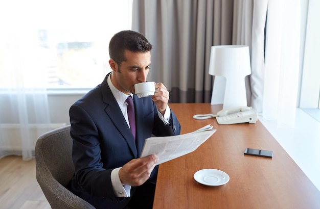 business trip, people and mass media concept - businessman reading newspaper and drinking coffee at hotel room