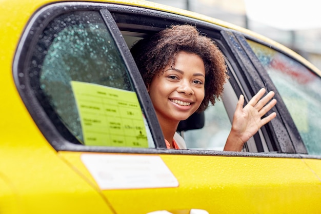 business trip, transportation, travel, gesture and people concept - young smiling african american woman driving in taxi and waving hand at city street