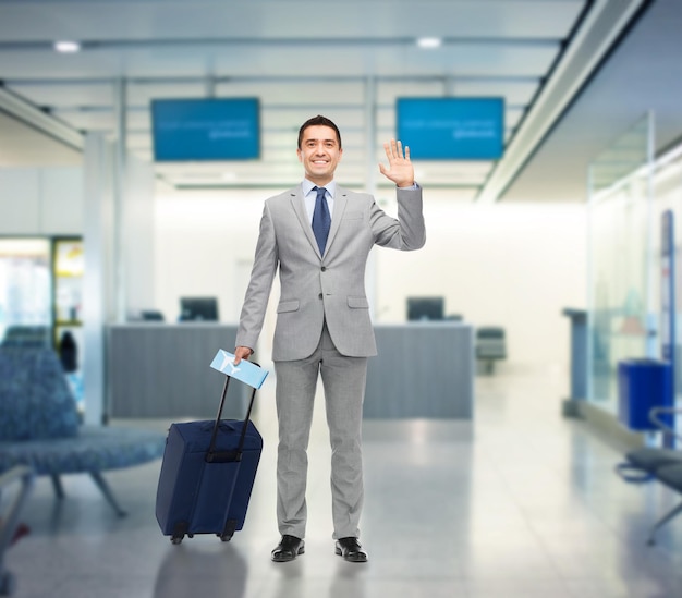 Концепция деловой поездки, путешествий, багажа и людей - счастливый бизнесмен в костюме с дорожной сумкой и авиабилетом, машущий рукой на фоне аэропорта