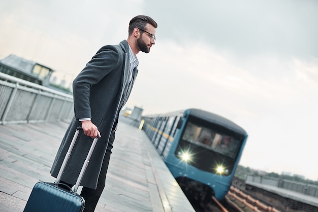 電車を待っている荷物を持って鉄道の近くに立っている出張青年実業家