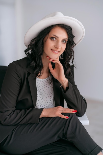 Business woman in a black suit and white hat sits on a cross and poses for a photo