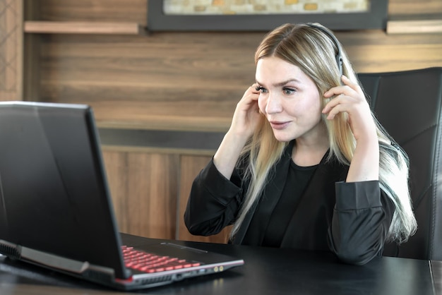 Business woman in black suit with headphones is sitting in front of laptop in office