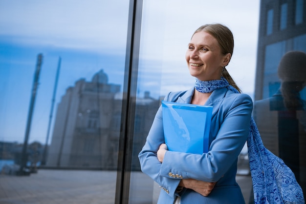 A business woman in a blue jacket holds a folder with papers in her hand in front of a glass office building concept of successful women