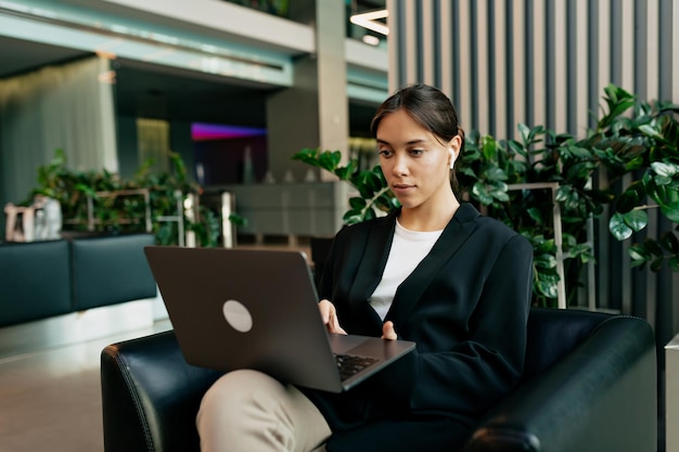 Business woman in dark jacket and white tshirt typing on laptop and chatting online with colleges with smile while sitting in her office