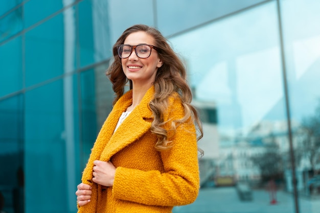 Business woman dressed yellow coat standing outdoors corporative building background Caucasian female business person eyeglasses on city street near office building big windows Stylish businesswoman