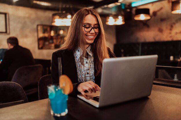 Business woman in glasses sitting in a modern cafe and working on her laptop while drinking a cocktail Selective focus High quality photo