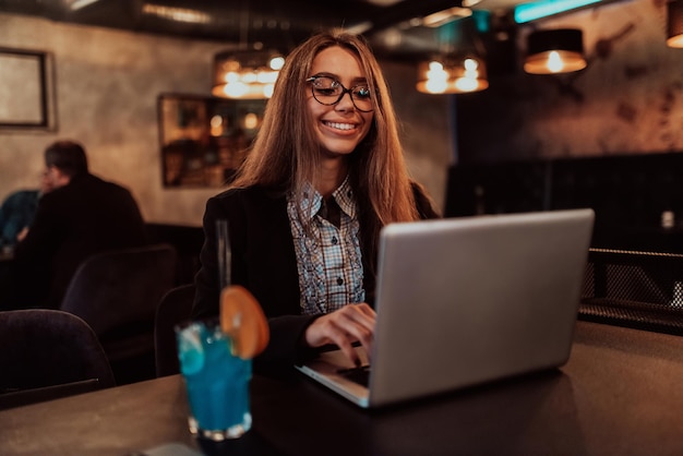 Business woman in glasses sitting in a modern cafe and working on her laptop while drinking a cocktail Selective focus