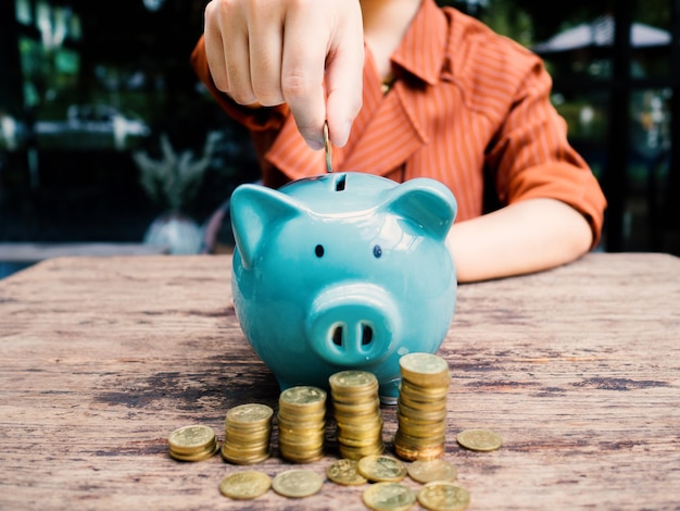 Business woman hand putting coin into  blue piggy bank with 
gold coins pile growth graph, Saving money for future investment plan and retirement fund concept.