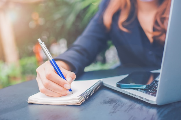 Foto scrittura della mano della donna di affari su un blocco note con una penna nell'ufficio. sul tavolo c'era un telefono cellulare e un computer portatile.