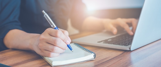 Foto mano della donna di affari che scrive su un blocco note con una penna e usa un computer portatile nell'ufficio.per il banner web.