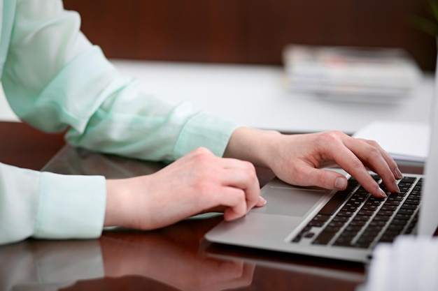 Руки деловой женщины в зеленой блузке сидят за столом в офисе и печатают на ноутбуке.