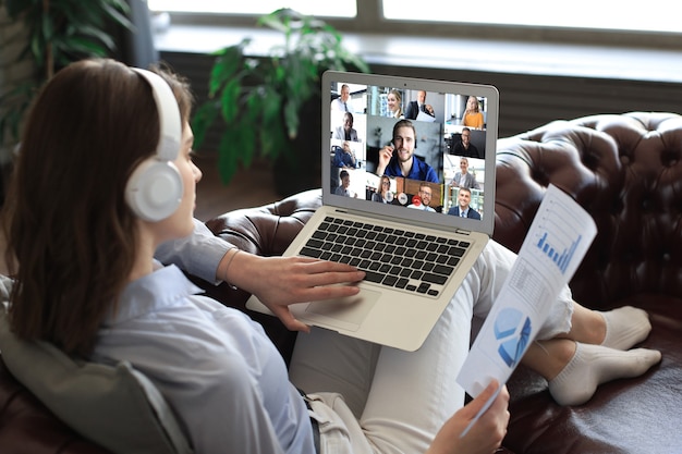 Business woman in headphones lying on sofa speak talking to her colleagues in video conference