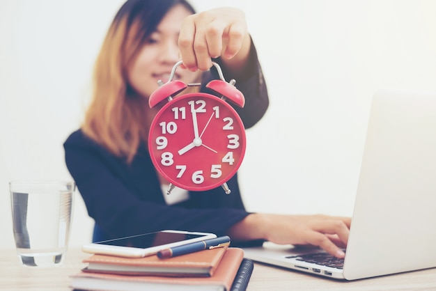 Photo business woman holding a red clock shows the time and a notebook with a laptop on the desk in the morning.