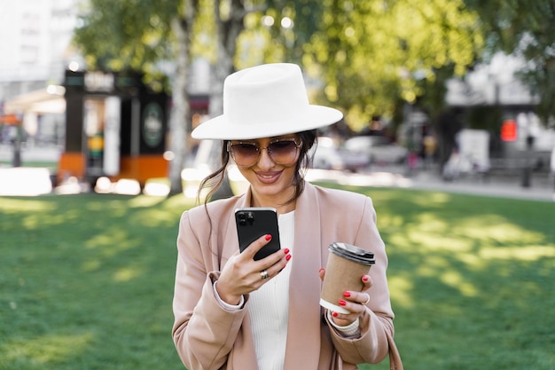 La donna d'affari sta chattando con gli amici per telefono e beve un caffè per strada. ritratto di stile di vita del modello di moda.