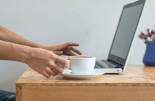 ビジネスの女性は、オフィステーブル上のラップトップPCで仕事中に白いコーヒーカップを保持しています