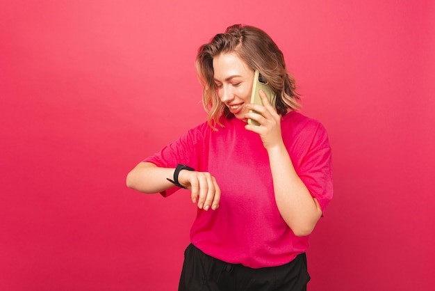 Business woman is talking to her phone while she is looking at watch near a pink wall