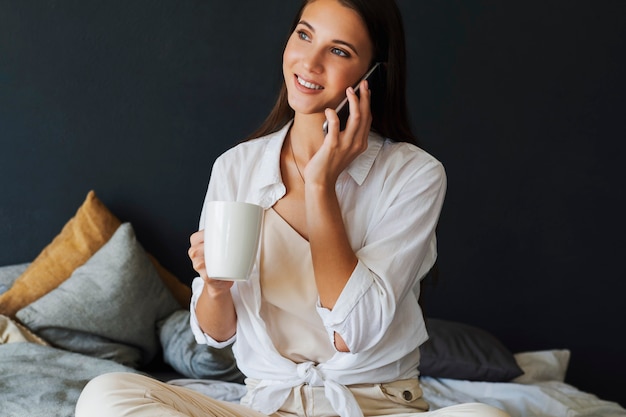 Business woman is talking on phone, negotiating, making an appointment with friends. Smiling girl in white shirt sits on bed, next to laptop. Brunette girl in the bedroom.