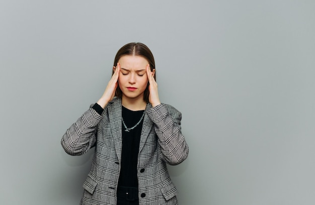 Business woman in a jacket stands on a gray background and suffers with a headache closes her eyes