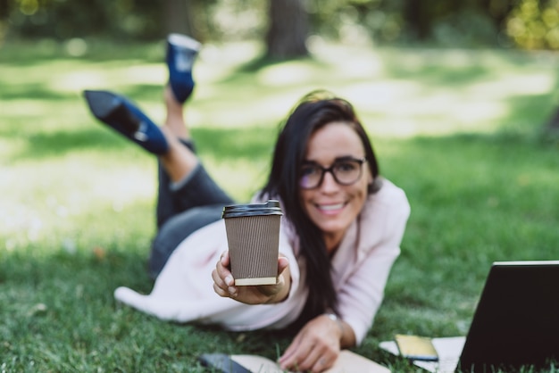 Business woman, lies in a summer grass park, using a laptop smiles and holds out a glass with coffee.