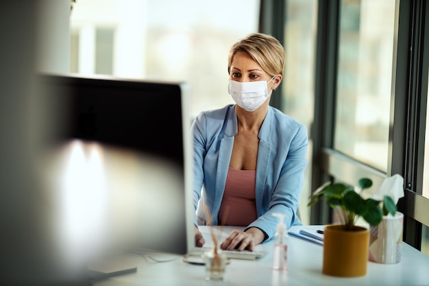 Business woman in a medical protective mask works at the office on the computer during self-isolation and quarantine to avoid infection during flu virus outbreak and coronavirus epidemic.