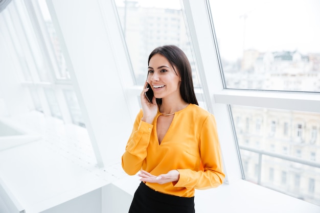 Photo business woman in orange shirt standing near the window and talking at phone in office