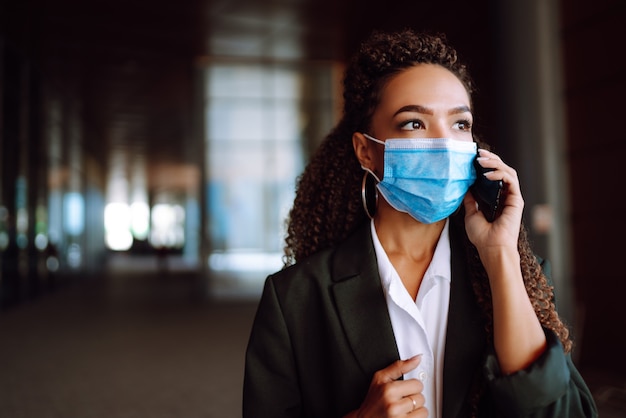 Деловая женщина в защитной медицинской маске, стоящей возле офиса и использующей телефон. COVID-19.