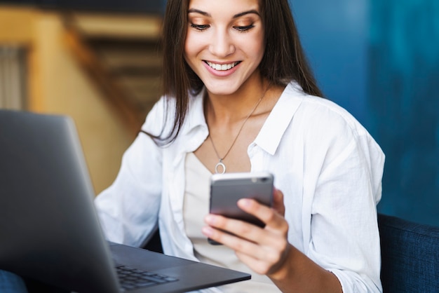 Business woman sits at table in front of laptop, writes message using smartphone, conducts business correspondence.