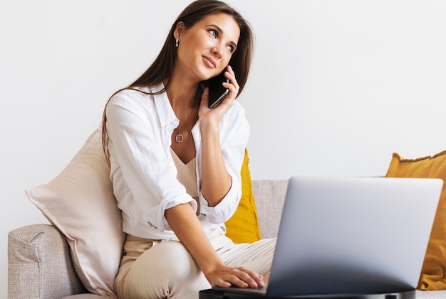 Business woman sits at table in front of laptop, writes message using smartphone, conducts business correspondence.