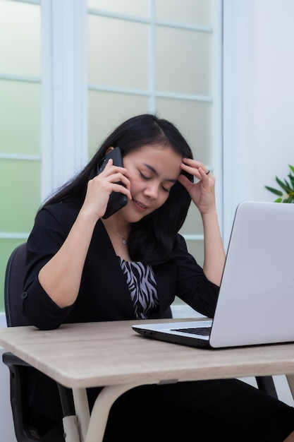 Business woman sitting on chair while making phone holding head in front of laptop