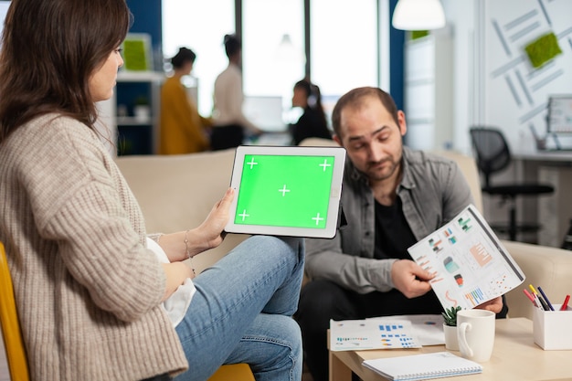 금융 통계를 분석하는 소파에 앉아 비즈니스 우먼, 다양한 팀이 배경에서 작업하는 동안 녹색 화면으로 태블릿을 들고
