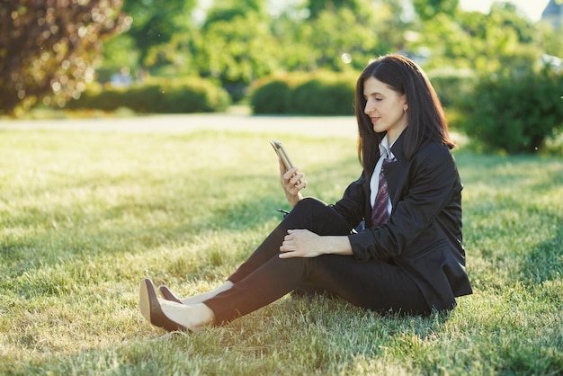 Business woman sitting on grass in sunny park with mobile phone in hand