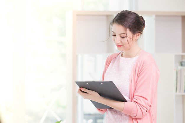 Business woman standing working near a window and holding clipboard at office.