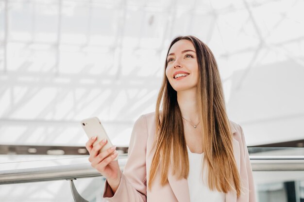 ビジネスの女性はオフィスビルの屋内に立ち、携帯電話を持って微笑んでいる