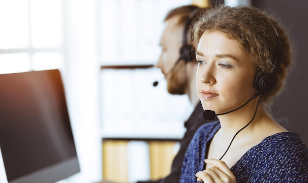 Business woman talking by headset while sitting with red-bearded colleague in modern sunny office. Diverse people group in call center.