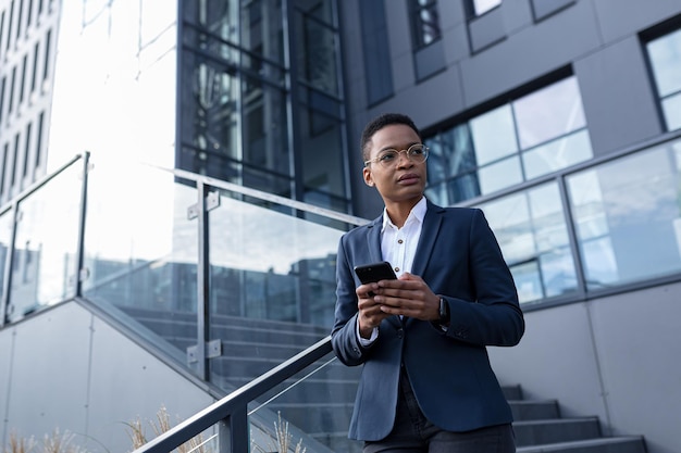 Business woman using cellphone, serious african american worker outside office